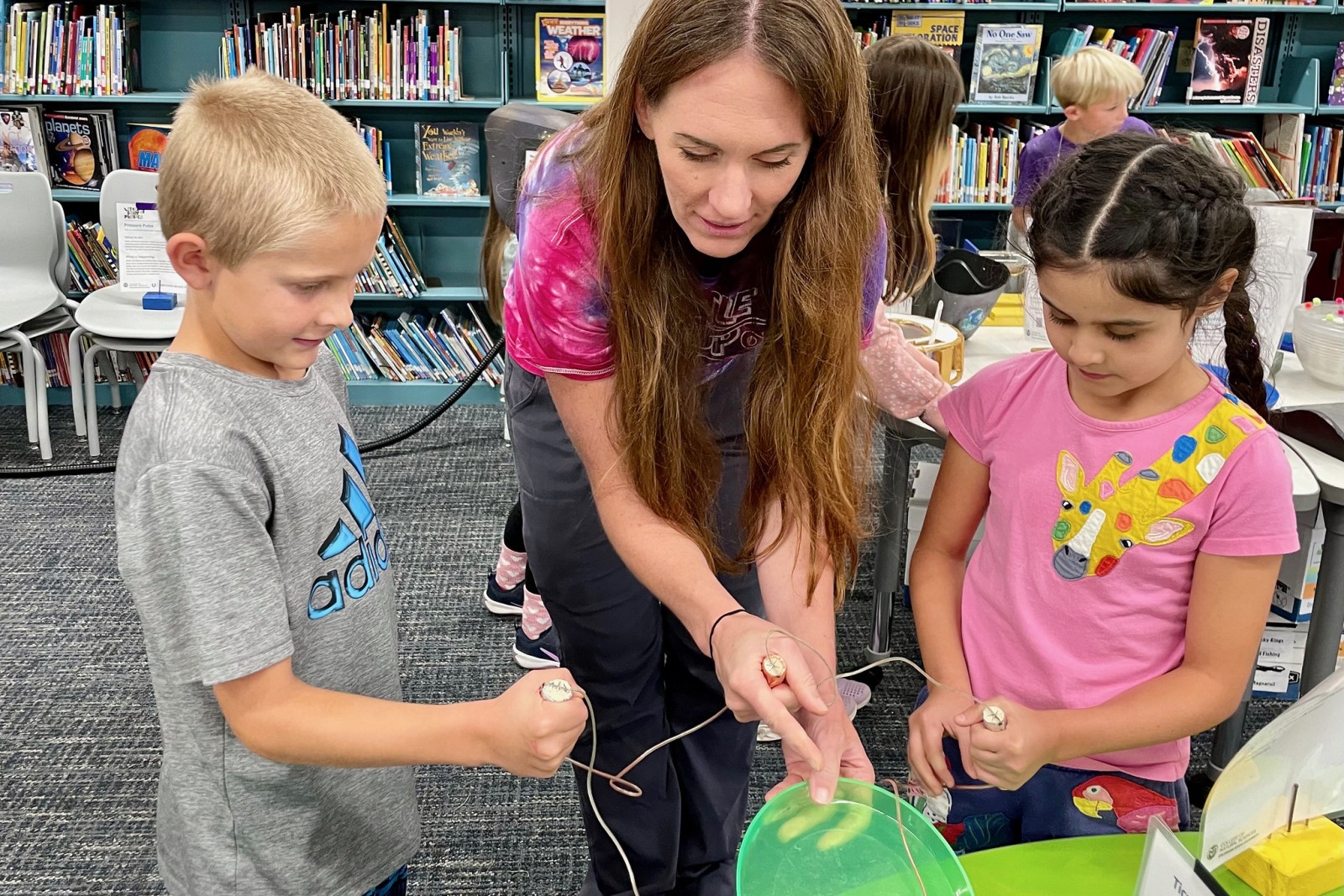 An adult stands between a boy and a girl elementary age, assisting with an science activity.