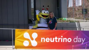 A bee mascot and a women stand behind a Neutrino Day banner.